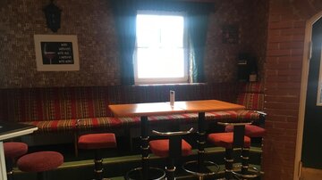 Ein gemütlicher Barbereich mit roten Hockern und einem Tisch. Im Hintergrund gibt es eine dekorative Wand und ein Fenster mit Tageslicht.