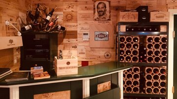 Ein gemütlicher Weinkeller mit Holzwänden, die Weinkisten zeigen. Im Hintergrund steht ein Regal mit Weinflaschen und ein Kühlschrank.