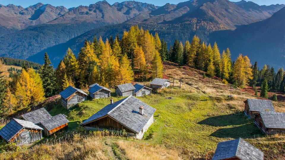 Eine malerische Berglandschaft mit mehreren Hütten und buntem Herbstwald. Im Hintergrund steigen majestätische Berge auf.