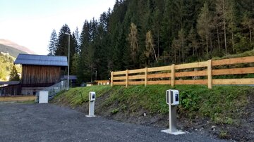 Eine ländliche Umgebung mit einem Holzgebäude und einer Zaunreihe. Im Vordergrund befinden sich zwei Ladestationen für Elektrofahrzeuge.