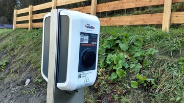 Eine Ladestation für Elektrofahrzeuge steht neben einer grünen Wiese. Im Hintergrund ist ein Holzzaun und Bäume zu sehen.