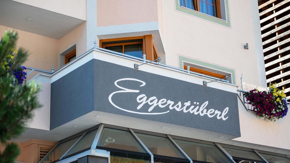 Ein modernes Gebäude mit einer Strickfront und einem großen Schild mit der Aufschrift „Eggerstüberl“. Blumen schmücken die Fenster und verleihen dem Ort eine einladende Atmosphäre.