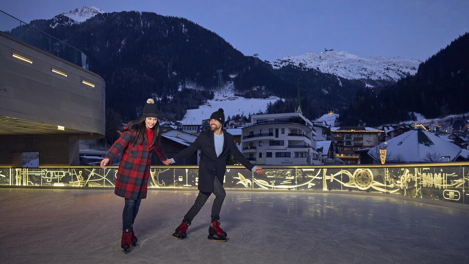 A couple is enjoying a lively round on an ice rink in the mountains. In the background, snow-covered peaks and cozy houses can be seen.