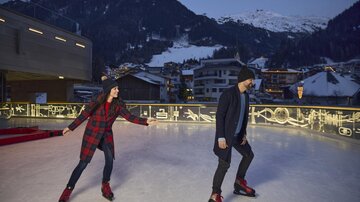Two people are skating on an ice rink at dusk. In the background, there are snow-covered mountains and cozy houses.