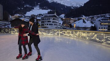A couple stands on an ice rink and enjoys skating. In the background, snow-covered mountains and winter buildings are visible.