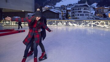 An ice rink with cheerful people having fun together. In the background, snow-covered mountains and winter houses are visible.