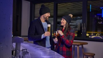 A man and a woman are standing at a bar and toasting with their drinks. Both are wearing hats and smiling at each other.