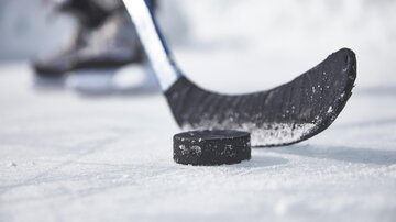 A hockey stick is standing on the ice next to a black puck. Blurred hockey players can be seen in the background. | © TVB Paznaun -  Ischgl