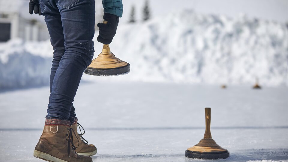 A person is standing on a frozen surface holding a wooden block in their hand. Snow-covered hills are visible in the background.