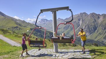 A summer toboggan run with two seating areas on a mountain meadow. In the background, green mountains and a blue sky can be seen.