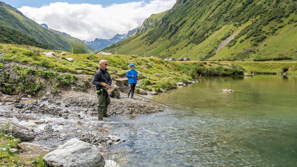 Zwei Angler stehen am Ufer eines klaren Gewässers und fischen. Die Umgebung ist grün und einladend.