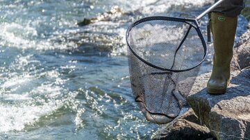 A person stands at the edge of a body of water with a net in hand. The water flows calmly and clear.