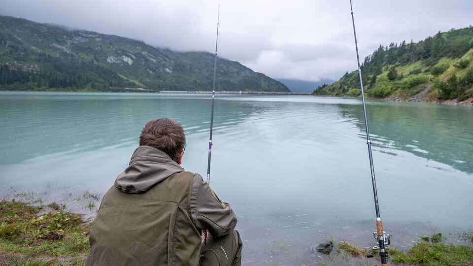 Two anglers stand by the shore of a clear mountain lake. Surrounded by green mountains and blue sky, they enjoy nature.