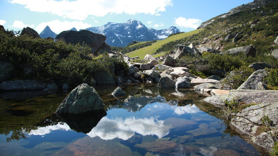 A picturesque mountain landscape with high peaks and a clear blue sky. In the foreground, the terrain is reflected in the calm water of a small pond.