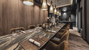 Ein eleganter Speisesaal mit einem langen Tisch und bequemen Stühlen. Die stimmungsvolle Beleuchtung und die holzverkleideten Wände schaffen eine gemütliche Atmosphäre.