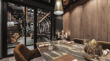 Ein elegantes Restaurant mit einem stilvollen Tisch und gemütlichen Stühlen. Im Hintergrund ist ein Weinschrank mit vielen Flaschen sichtbar.