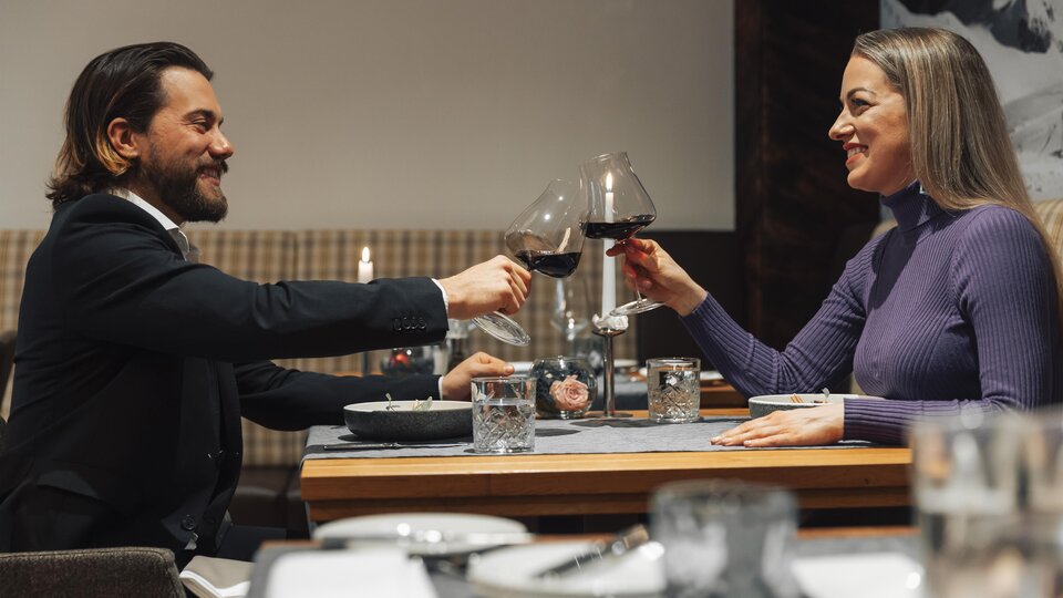 Ein elegantes Dinner-Paar stoßen mit Rotwein an. Der Tisch ist gedeckt und die Atmosphäre wirkt gemütlich.