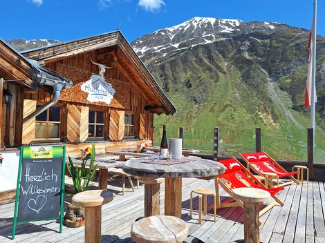 Eine gemütliche Berghütte mit Holzelementen und einer herrlichen Berglandschaft im Hintergrund. Es gibt draußen ein Publikum mit Liegestühlen und ein Willkommensschild.