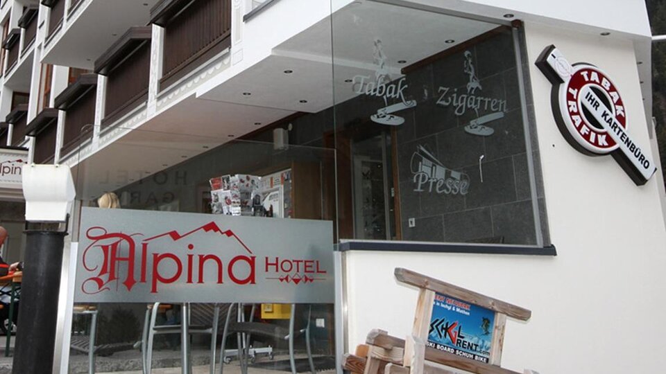 A modern hotel with the sign "Alpina Hotel" is visible. The facade has large windows and features an inviting entrance area.