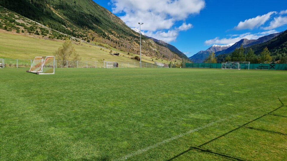 Ein grünes Fußballfeld in einer malerischen Berglandschaft. Im Hintergrund sind hohe Berge und ein klarer blauer Himmel zu sehen.