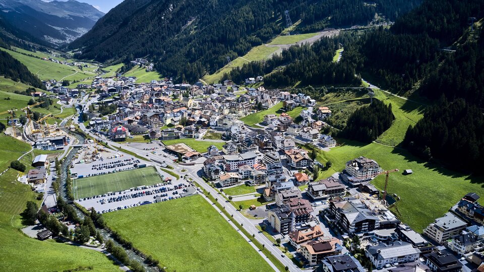 Eine malerische alpine Stadt in einem grünen Tal, umgeben von Bergen. Die Gebäude sind dicht gepackt, und es gibt viele Parkplätze und Straßen.