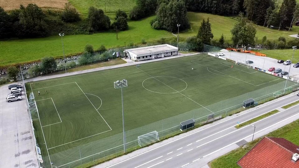 A sports field with artificial turf, surrounded by trees. In the background, a building and parked cars are visible.