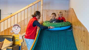 A room with a slide on which two children are sitting. A caregiver is helping them slide down.