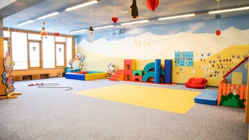 A colorful playroom with play equipment and soft flooring. The walls are decorated with child-friendly motifs.