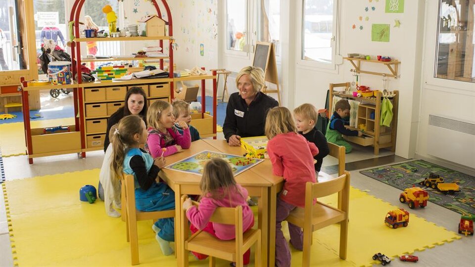Ein heller Kindergartenraum mit Kindern, die um einen Tisch sitzen und spielen. Eine Betreuerin ist anwesend, während im Hintergrund weitere Kinder mit Spielzeug beschäftigt sind.