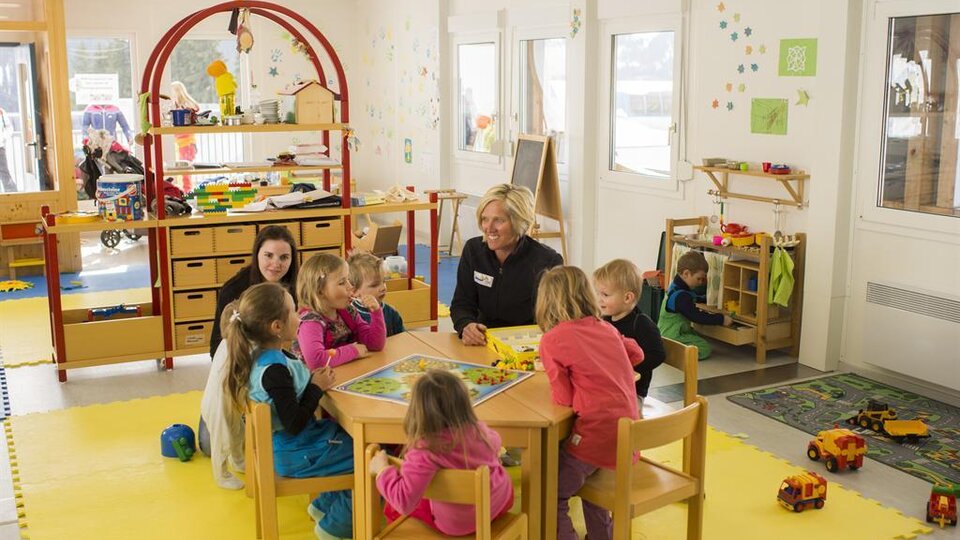 A bright kindergarten room with children sitting around a table and playing. A caregiver is present, while in the background, other children are occupied with toys.