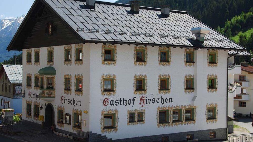 Ein traditionelles Gasthaus mit der Aufschrift „Gasthof Hirschen“ an der Fassade. Das Gebäude hat eine historische Architektur und befindet sich in einer malerischen Berglandschaft.
