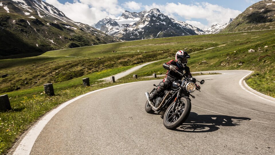 A motorcyclist rides on a winding road in the mountains. The landscape is green with snow-capped peaks in the background.