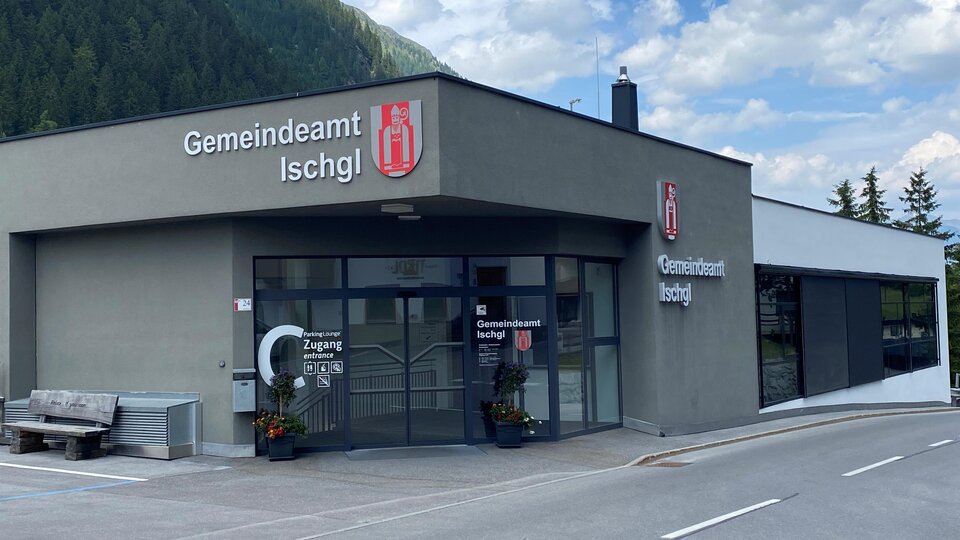 A modern building of the Ischgl municipal office with clear signage. In the background, green mountains and a blue sky can be seen.