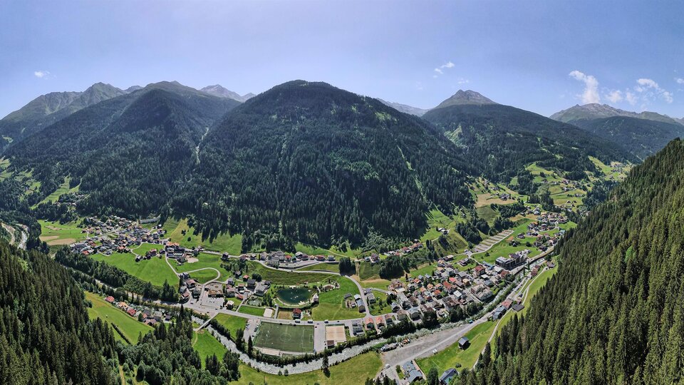 Ein schöner panaromatischer Blick auf ein Tal umgeben von hohen Bergen. Die Landschaft ist grün mit Wäldern und einem kleinen Ort im Hintergrund.