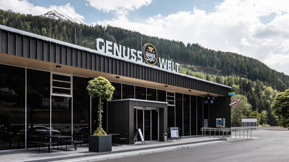 A modern building with the inscription "GENUSS WELT" stands in a green landscape. In the background, mountains and a clear sky are visible.