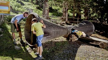 Two children and an adult are exploring a large horn in nature. Trees and a green landscape can be seen in the background.
