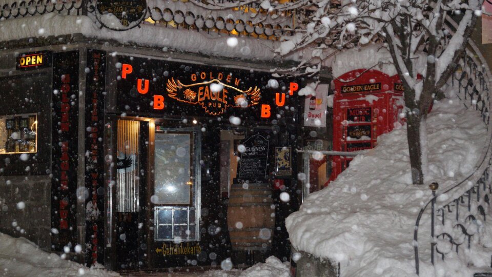 Ein gemütlicher Pub mit dem Namen "Golden Eagle" ist im Schnee verborgen. Die Schneeflocken fallen dicht, während das warme Licht des Eingangs einladend scheint.