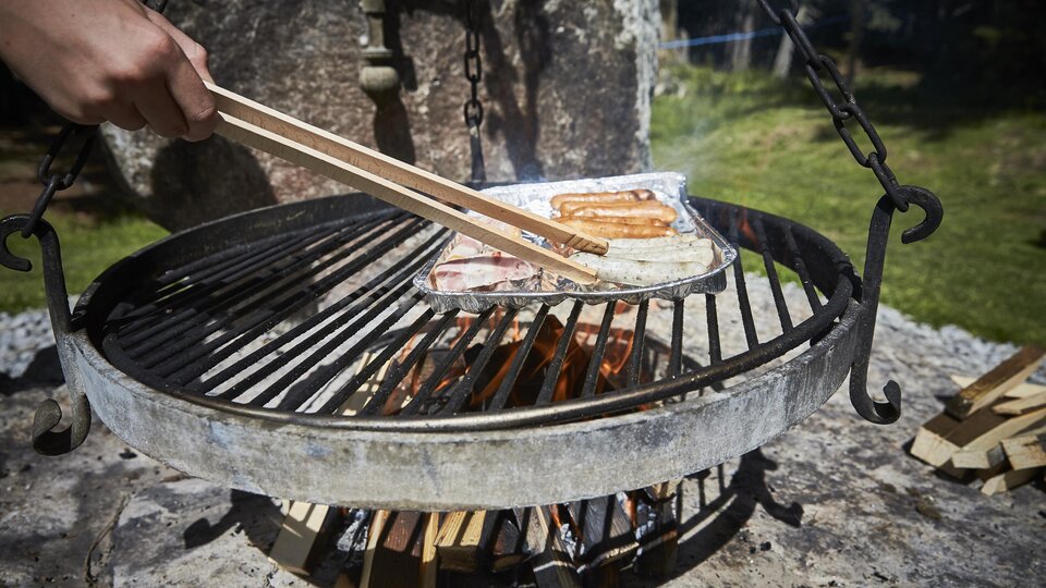 A grill with sizzling sausages is being prepared over an open flame. In the background, green grass and nature can be seen.