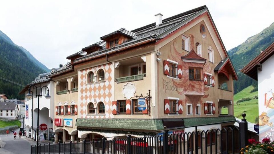 Ein charmantes Gebäude mit kunstvollen Wandmalereien in einer malerischen Berglandschaft. Die Straßen sind von Natur umgeben, und die Atmosphäre ist einladend.
