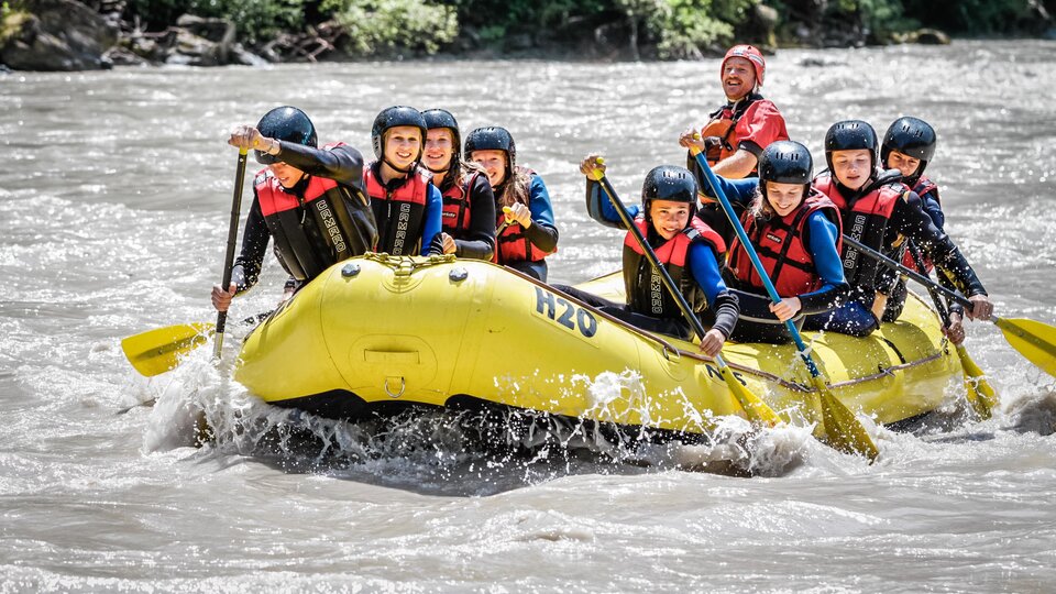 Eine Gruppe von acht Personen paddelt in einem gelben Raft auf einem Fluss. Sie tragen Helme und Schwimmwesten und haben viel Spaß.