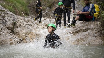 Eine Gruppe von Kindern und Erwachsenen hat Spaß beim Wassersport in einem Fluss. Einige tragen grünes Sicherheitshelme und spielen im Wasser.