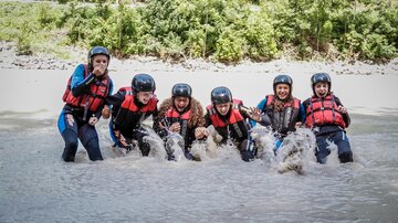 Eine Gruppe von sechs Personen steht im Wasser und hat viel Spaß. Alle tragen Schwimmwesten und Helme.