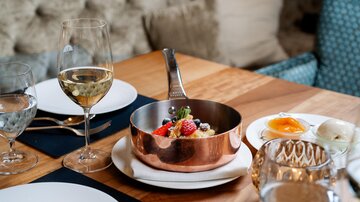 Ein elegant gedeckter Tisch mit Gläsern, einem copperfarbenen Topf voller frischer Früchte und einer weißen Sauce. Ein ruhiges Ambiente mit geschmackvoller Dekoration.