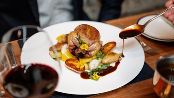 Ein köstliches Gericht mit Brathähnchen, Gemüse und Sauce auf einem weißen Teller. Im Hintergrund sind Wein und eine elegant gedeckte Tafel zu sehen.
