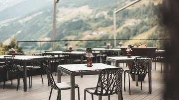 A terrace with several tables and chairs, surrounded by mountain landscape. The atmosphere is inviting and calm.