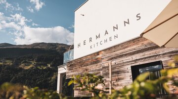 A modern restaurant facade with the inscription "Hermann's Kitchen". In the background, mountains and a blue sky are visible.