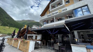 Ein gemütliches Hotel mit Außenbereich und Terrassenmöbeln. Im Hintergrund sind grüne Berge und ein bewölkter Himmel sichtbar.