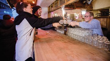 Eine lebhafte Bar mit fröhlichen Gästen, die Getränke bestellen. Ein Bartender serviert freundlich, während die Atmosphäre gemütlich ist.