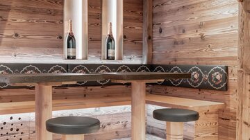 Eine gemütliche Holzbar mit zwei Hockern und einer dicken Tischfläche. Im Hintergrund stehen zwei Flaschen Sekt in eleganten Halterungen.