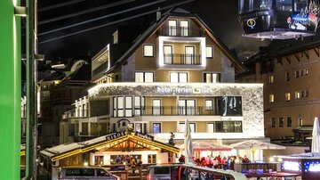 Ein modernes Hotel mit beleuchteter Fassade in einer Alpenstadt bei Nacht. Neben dem Hotel sind ein Souvenirstand und Menschen, die sich amüsieren.
